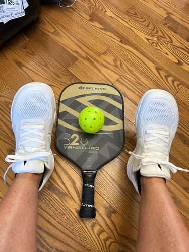 Customer's white Cuurt Muuv court shoes on feet with a pickleball paddle and ball on a wooden floor.