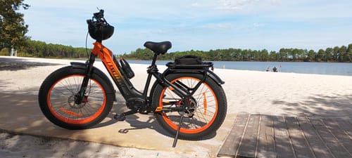 Customer's orange and black Lankeleisi MG600 Plus fat bike parked on a wooden path near a sandy beach.
