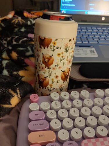 Customer's Highland Cow Universal Buddy can cooler with floral pattern, sitting on a blanket near a laptop and keyboard.