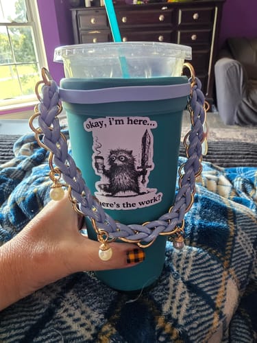 Customer's hand holding a teal Frost Buddy cup with a decorative braided handle and a raccoon sticker, on a patterned rug.