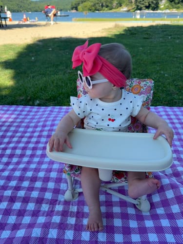 Reviewer's floral print Paloma portable booster chair V2 with tray, set on a purple checkered picnic blanket outdoors.