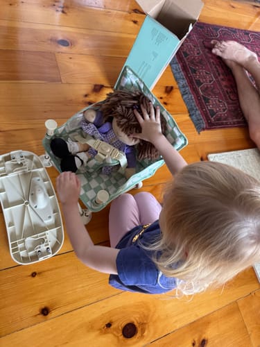 Reviewer's child placing a hand on a doll inside the checkered Parker portable booster chair V2 on a wooden floor.
