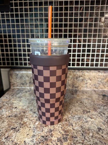 Reviewer's Espresso Checkered To-Go Buddy cup insulator holding an iced drink with a straw on a kitchen counter.