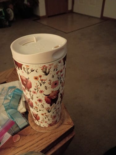 Customer's To-Go Buddy cup with a highland cow pattern, featuring a light-colored lid, resting on a wooden surface.