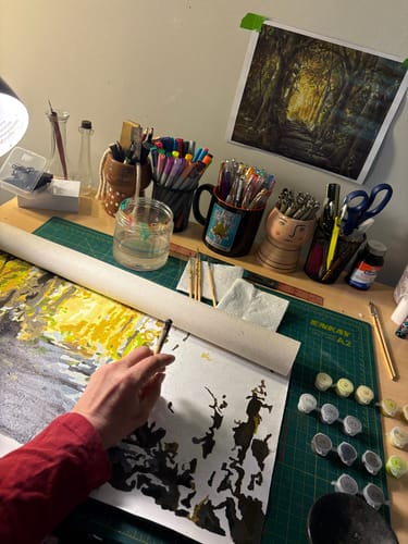 Customer's hand holding a Silver OG High Grade Pre Roll while painting on a workspace desk with art supplies.