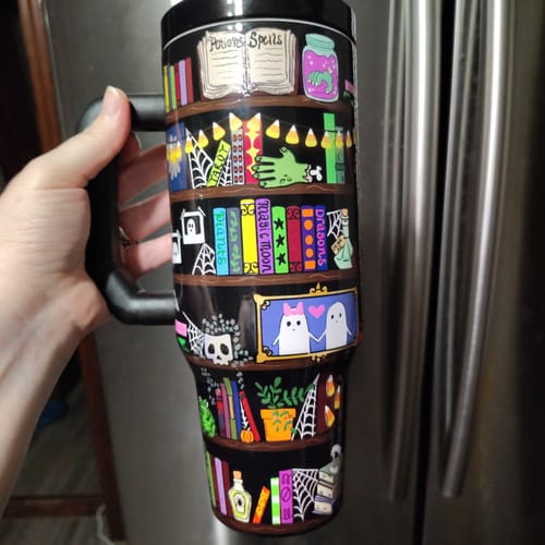 Customer's Thicc Buddy mug held in hand, showing vibrant bookshelf illustration with potions, ghosts, and spiders.