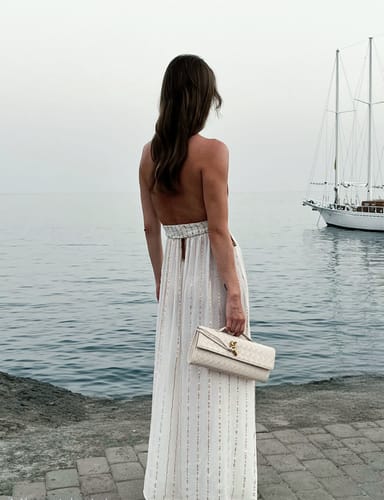 Customer's cream Bianca Woven Bag, held by a person in a dress near the water.