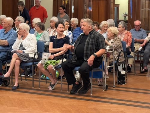 Reviewer's gathering of people sitting in a room, the reviewer is wearing a bright, floral, asymmetrical-hem top.