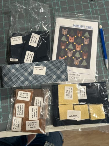 Customer's Midnight Pines quilt kit, showing pre-cut fabric bags, instruction booklet, and binding fabric on a desk.