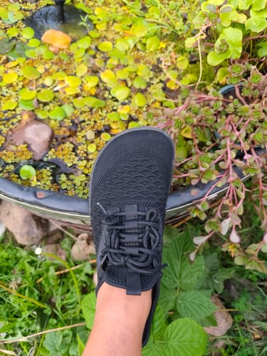 Customer's black Go I Barfußschuh worn in a natural garden setting near a small pond.
