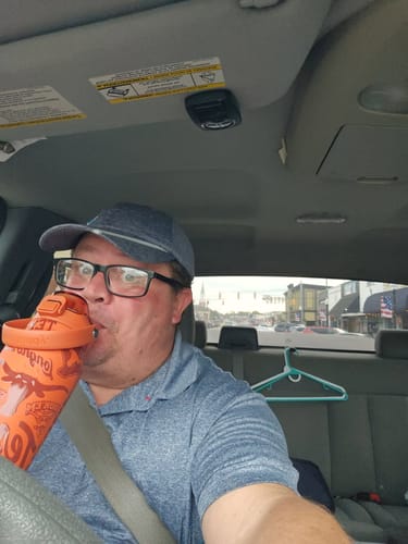 Reviewer in a vehicle drinking from an orange Texas Longhorns Bottle Buddy 32oz while wearing a cap.