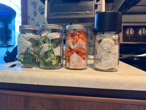 Reviewer's Mason Genie Vacuum Sealer Kit used to seal four glass jars filled with vegetables on a kitchen counter.