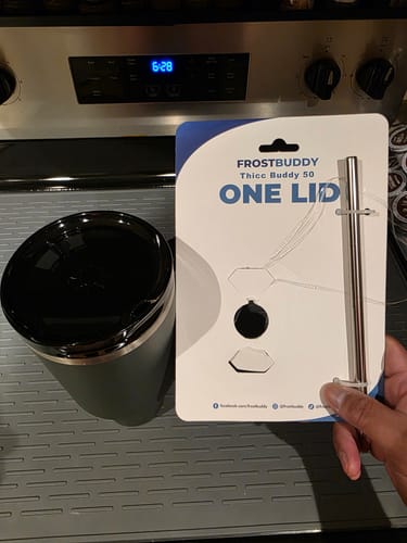 Customer's ONE Lid packaging for Thicc Buddy held by a hand next to a dark gray cup on a kitchen stove.