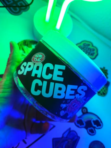 Reviewer's hand holding a round container of THC Space Cubes under bright green light.
