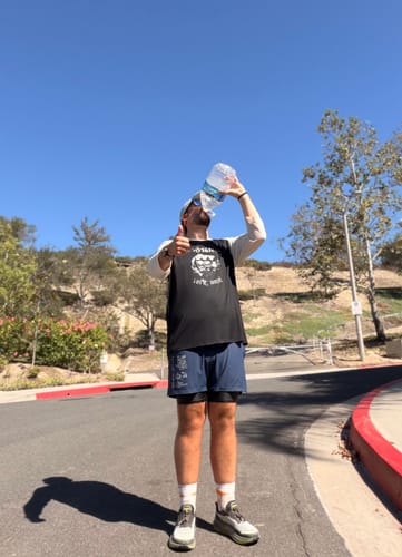 Customer's PlayOn Pain Relief Spray, held by runner giving a thumbs up while drinking water outdoors.