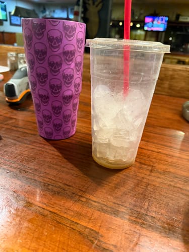 Customer's purple To-Go Buddy tumbler with a skull pattern sitting next to a plastic cup with ice on a wooden table.