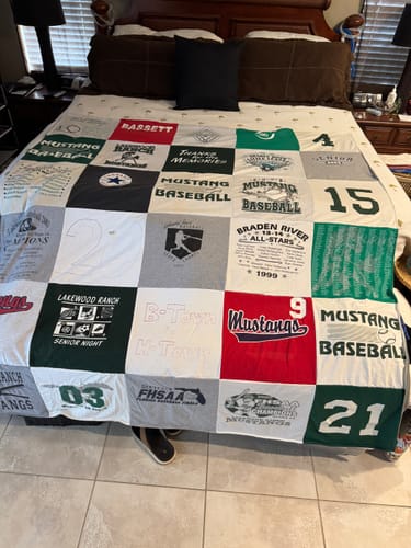 Customer's Project Repat T-Shirt Quilt displayed on a bed, showing various custom athletic logos and team numbers.