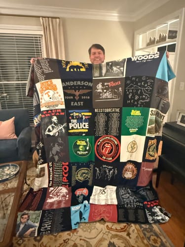 Customer's Project Repat T-Shirt Quilt held up in a living room, featuring a collection of band and concert t-shirts.