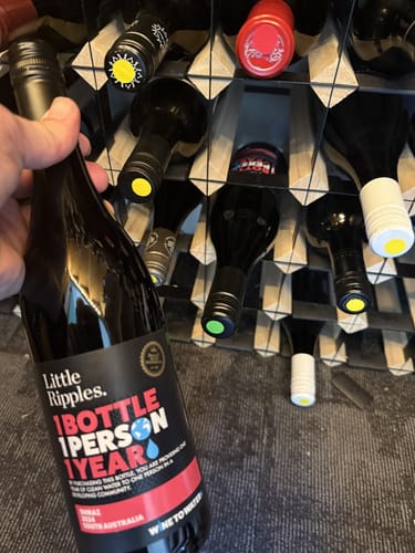 Customer's hand holding a bottle of Little Ripples wine in front of a wine rack with various bottles