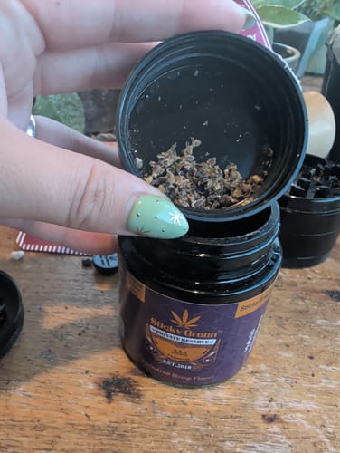 Reviewer's hand holding open black jar of Royal Runtz flower, showing a small amount of ground flower inside the lid.