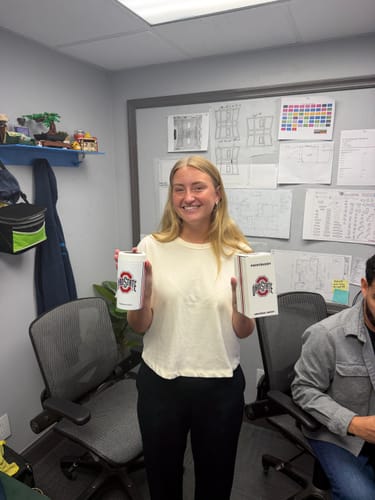 Customer's smiling coworker holding the Ohio State Universal Buddy can cooler and its white packaging in an office.