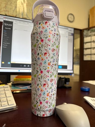 Reviewer's Bottle Buddy in Wildflower print standing on a wooden desk next to a computer mouse and keyboard.