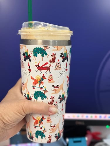 Customer's hand holding a white Frost Buddy cup patterned with colorful holiday dogs.