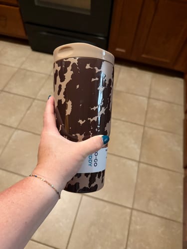 Customer's hand holding a brown and tan cow-print To-Go Buddy tumbler with the product label still attached.