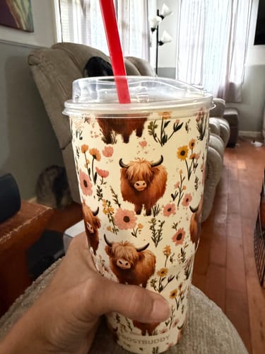 Customer's hand holding a cream-colored To-Go Buddy tumbler with a highland cow and floral pattern, featuring a red straw.
