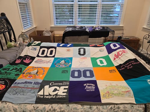 Customer's Project Repat T-Shirt Quilt displayed on a bed, showcasing a colorful patchwork of various sports logos.