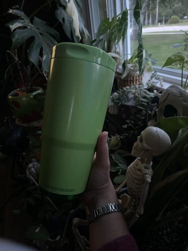 Customer's lime green To-Go Buddy tumbler held in a hand, surrounded by indoor plants and a decorative skull.