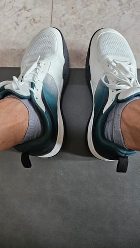 Customer's feet wearing white and blue CUURT Muuv court shoes while standing on a gray surface.