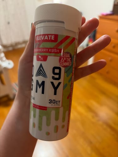 Reviewer's hand holding a jar of Strawberry Kush Delta 9 Gummies in a home setting.