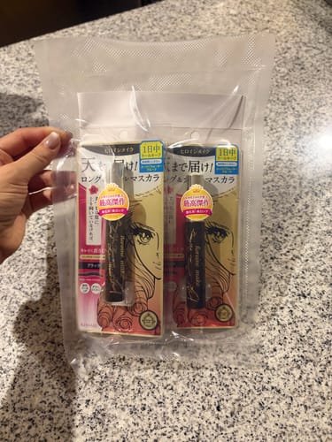 Customer's two Kiss Me Heroine Mascara tubes sealed inside a clear plastic vacuum bag, held by a hand.