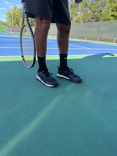 Customer's black CUURT Muuv court shoes worn while holding a tennis racket on a blue tennis court.