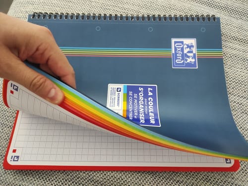 Reviewer's hand lifting the blue cover of a spiral-bound Oxford Color System notebook, revealing rainbow-colored pages.