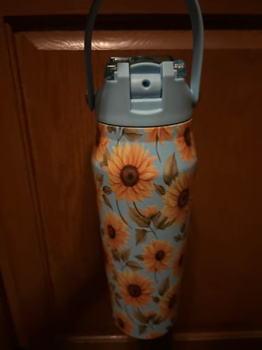 Customer's Bottle Buddy water bottle with blue lid and sunflower pattern against a wooden cabinet.