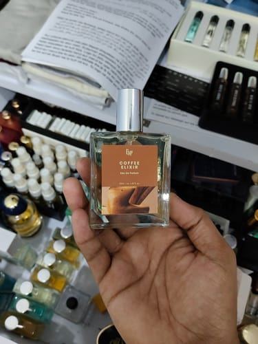 Reviewer's hand holding a clear, square Coffee Elixir bottle with a brown label among many other fragrance bottles.