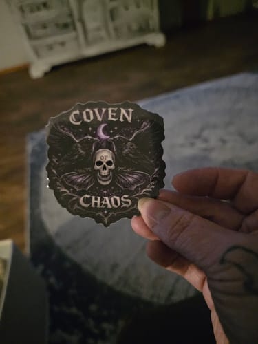 Reviewer's hand holding a round 3-inch Coven of Chaos sticker with a skull, ravens, and moon design.
