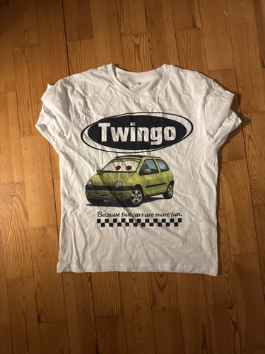 Kunde zeigt sein weißes personalisiertes T-Shirt mit einem lustigen Aufdruck eines grünen Twingo-Autos auf einem Holzboden.