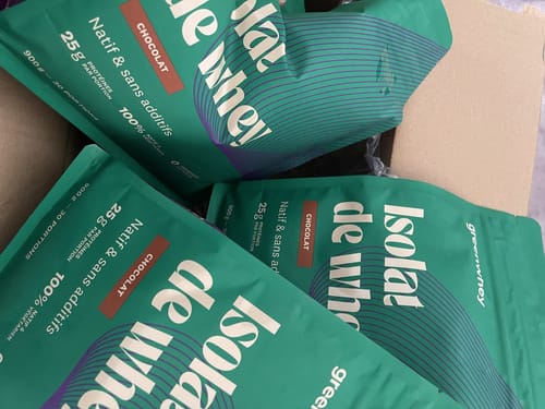 Photo d'un client montrant plusieurs sachets verts d'Isolat de Whey Native goût chocolat dans un carton.