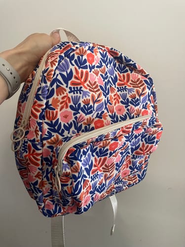 A customer holds up the floral Willow backpack, showing the main compartment and front zippered pocket.
