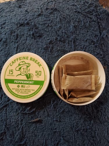 Customer's open round tin of Peppermint Caffeine Pouches next to its lid, showing the brown pouches inside.