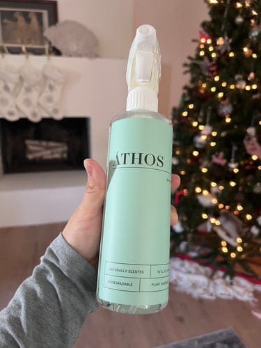 Customer holding a spray bottle of ÁTHOS cleaner with a mint-green label in their home.