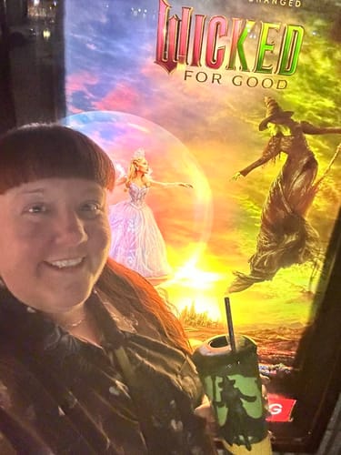 Reviewer wearing the dark, patterned "Wicked Winds" shirt while posing in front of a large movie poster for "Wicked."