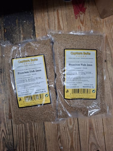 Klant toont twee doorzichtige zakken Captura Baits Premium Fish 2mm pellets op een houten ondergrond.