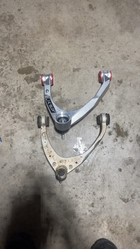 Customer's new KSP aluminum control arm shown above the old stock part for comparison on a concrete floor.