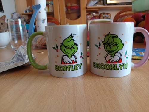 Kunde zeigt zwei personalisierte Grinch-Tassen mit den Namen „Bentley“ und „Brooklyn“ auf einem Tisch.