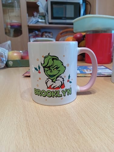 Kunde zeigt die personalisierte Grinch-Tasse mit dem Namen Brooklyn und einem rosa Henkel auf einem Holztisch.