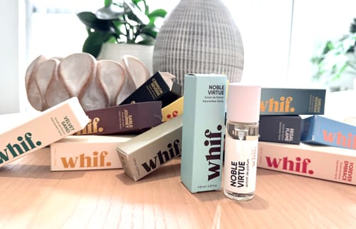 Reviewer's small Noble Virtue perfume bottle and its box on a wooden table among a collection of other colorful fragrances.